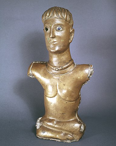 A gleaming bronze statue of a young man with short-cropped hair and a torc. He has naturalistic but disproportionate features. His left eye is wide open with white and black glass / enamel, but the right eye is missing. His head looks over large for His body, and both arms are missing. Where you would expect His bellybutton is where His legs are crossed at, one lying fully horizontal and parallel over the other. His shoes make his feet look almost like hooves.
