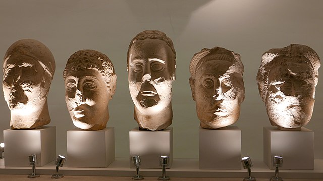 Five white stone heads mounted on museum pedestals. They have tall, somber faces with large almond shaped eyes, and round curving eyebrows. Their features are much more naturalistic than the Roquepertuse Janus and each has a unique hairstyle while one wears a helmet and two of them have what may be hats. All are partly damaged or mutilated.