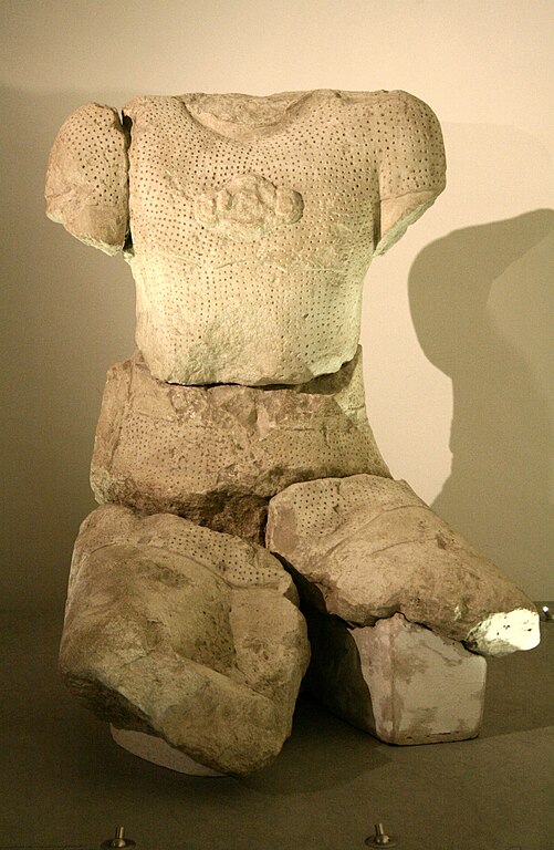 A cross-legged white stone statue similar to the Glanum Celt, also missing the head and both arms. This one has no restored paint, and has heavily damaged legs that are even more large and out of proportion. The torso wears a cuirass that has some sort of three-circle design over the chest. The clothes are textured with perforated holes all over. The different body parts have been reassembled after being broken apart.