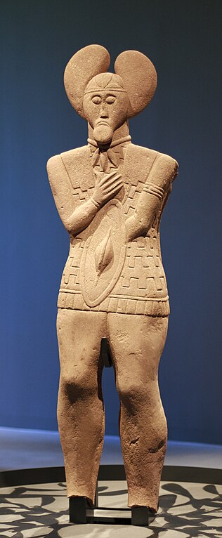 Photo of the Glauberg Prince, a well-preserved reddish-orange stone statue of a man holding a tiny shield. His free right hand folds over His chest and He has the so-called mistletoe leaf headdress. A horizontal line and array of leaf shapes marks His hair or headgear. He has large almond shaped eyes and a droopy frown with large chin or goatee. He wears a necklace from which three leaf shaped objects hang in front. He has an armband and a patterned tunic or armored cuirass with fringe. His legs are massive like trunks.