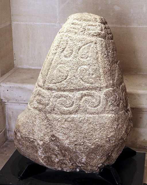 Photo of a large, conical gray stone with four sides. The side facing the viewer has a central panel with four tear-drop shapes in each corner facing away from each other in opposite directions. Below is a boarder of waves or swirling pattern, and above is a border with angular design perhaps like a labyrinth. The bottom third of the cone is left plain.