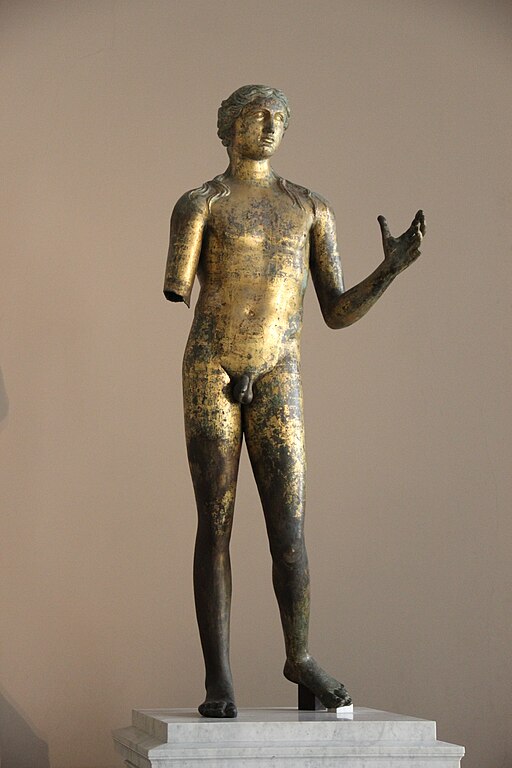 A large bronze statue of Apollo gilded gold, mounted on a polished white marble base. He is nude and has long, curly locks that drape over His shoulders. His is in contrapposto with His left foot lifted off the ground. He looks out, with His left hand stretched out, but His right arm is missing from the elbow down. He is sculpted in Classical style, albeit a bit stiff looking and lacking in finer details.