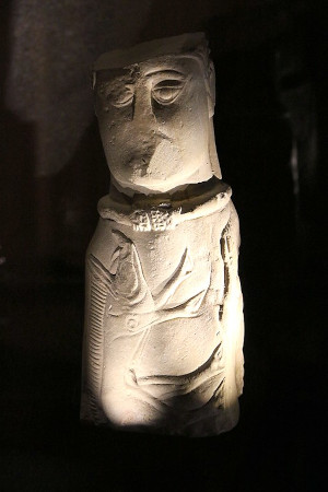 A photo of the Euffigneix bust, in the shape of a small white pillar, under a stark light that casts shadows on its low relief designs. It has a Celtic style boar emblazoned across the front torso. He wears an oversized torc. His face and scalp is mutilated, but one can still see His large, graceful almond shaped eyes with a gentle expression.