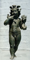 A small but life-like black bronze figurine of Harpocrates depicted as a plump toddler boy. He stands with one foot poised in front of the other and holds His right index finger up to His chin with a coy look on His face. He is naked except for some sort of headdress and a necklace that suspends a large, round ball. He holds a small cornucopia in His left hand and some indiscernable object sits on His right shoulder.