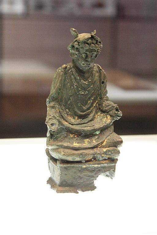 A photo of a cross-legged seated tarnished-bronze Mercury with curly hair Who wears the winged petasus on His head and a full toga. His arms rest on each knee. The statuette is well-preserved except that His hands are missing and a corner of the base that it's affixed to is broken off.