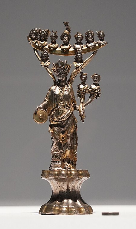 A gleaming silver figurine of Tutela with a row of busts mounted on Her wings. The heads represent the 7 Gods of the days of the week. Beneath Them are the heads of the Dioscuri. She holds a double cornucopia in Her left hand mounted by the busts of Apollo and Diana.