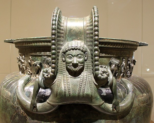 Photo of the top portion of an elaborate green-black bronze krater. The handle shows the bust of a grimacing Gorgon in Archaic Greek style. She has large almond shaped eyes and Her tongue sticking out. Two serpents stretch from beneath Her bosom to attach the bottom of the handle to the vase. Her arms are held outward and bent down at a ninety degree angle. The handle also stretches up behind Her head to attach to the top of the vase, with a patterned border. The neck of the vase is surrounded with a relief of soldiers and other decorative borders.