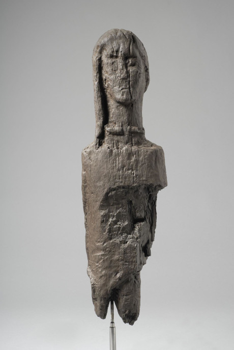 Partially damaged timber post suspended on a metal rod. It has a plastic looking texture. The top is sculpted as a tall, solemn looking woman's face. The details only faintly remain, with a crack running down Her face and neck through the corner of Her left eye. She has long hair and wears a torc.