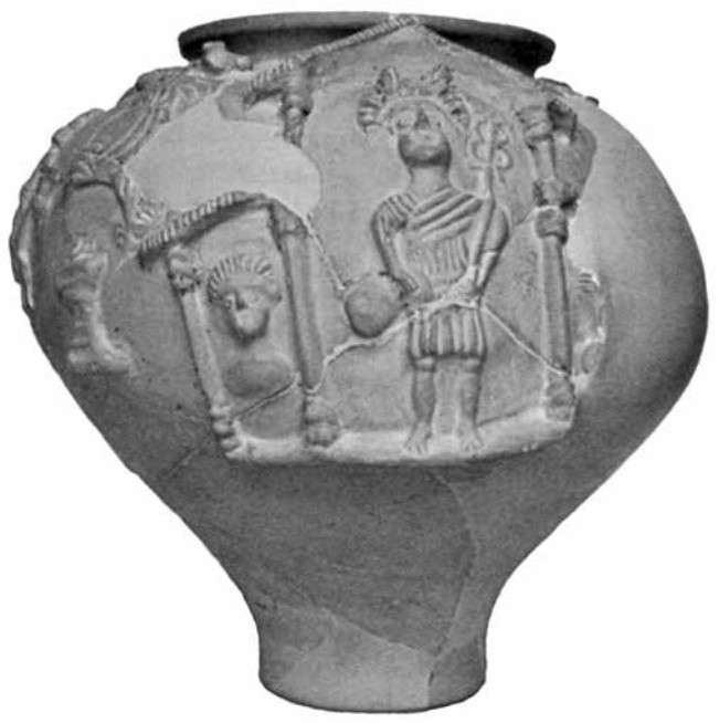 A black-and-white photo of a large, wide vase with a narrow base. On the front is a relief of Mercury standing under a roof with columns, beside a disembodied face that is also under a shorter roof, around half the height of Mercury. Mercury fills the full height of the building. He wears a tunic, has wings on His head, and holds His caduceus in His left hand, money bag in His right. The structure is assumed to be a cross-section depiction of a Gallo-Roman fanum, with the shorter roof representing the ambulatory that surrounds it.