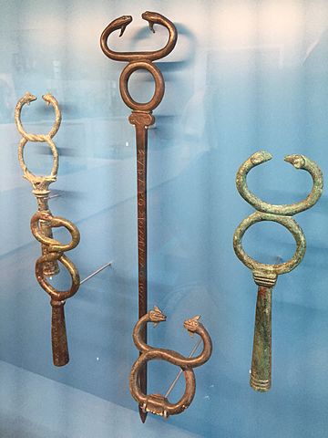 A glass museum case showing one full bronze kerykeion and the tops of 4 others. They each show the two snakes curling up into a figure-8, not quite meeting each other at the top for a kiss. Some of the figure-8s are actually twisted into shape, while others just welded the two snake heads on top of a ring