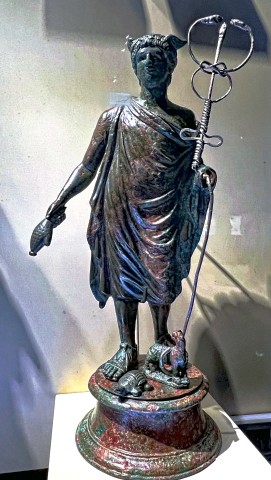 A dark bronze figurine of Mercury on a pedestal with a small tortoise and goat. He wears a full-length chlamys, holds a money bag in His right hand, and a caduceus in His left. The caduceus is a thin rod with a wire coiled around it from the ground up, forming both its wings and its snakes which tie in an elegant knot and nearly kiss at the top of their figure-8.