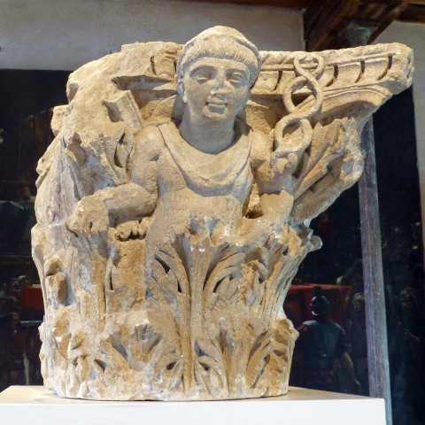 Photo of one side of a square, limestone Corinthian capital with the upper left corner smashed off. The upper body of the God Mercury emerges from the acanthus leaves below with a soft smile on His lips, large almond shaped eyes, short straight hair, and a small petasos on His head. His nose is broken off. His right hand is held out to the side with a faint remnant of what was probably a money bag. His left hand upholds the caduceus, which has no wings. Its two snakes coil in a double helix that forms three loops.
