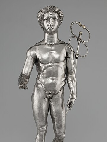 A polished silver statue of the God Mercury completely in the nude, reaching out His right hand, and holding the caduceus against His shoulder with the left. The caduceus is a slender rod with a ball on top. The snakes are silver wire that form an oblong figure-8. The wire twists around at both the base and in the middle where it forms an actual knot. The snake heads are small golden ovals at the ends of the wire that are closed together at the top, and faintly have the details of their faces.