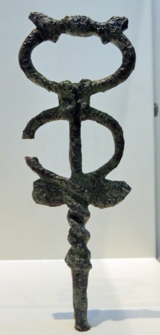 The top part of a mottled, damaged bronze caduceus on display in a museum. It has two oval rings that are bound together with a double wrap of the bronze tubing. The staff extends up through the bottom ring. At the base of the bottom ring is a pair of small wings, almost like a hummingbird's. The tubing that forms the bottom ring coils around the staff below.
