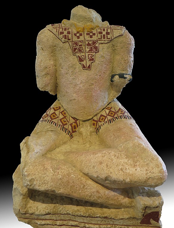 A headless white, stone, cross-legged figure with missing forearms that has been painted to restore some of the polychromy. A portion over the shoulders and chest as well as the short skirt around His upper thighs are painted with red and yellow mostly square, diamond, and triangle geometric patters. The skirt also has a fringe painted around the bottom. There is a dark bracelet around the left bicep. The statue's calves are disproportionately large and muscular. The torso has no detail to it. A large weathered phallus is pulled out over the top of the warrior's skirt.