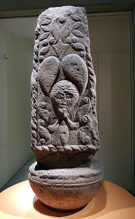 A gray stone rectangular pillar or stela almost in the shape of a buoy for its hemispherical base that it balances on. On the front is the head of the Glauberg Prince, virtually identical down to each detail, except for the necklace which is styled more like the neck ruffle of a jester. The head is surrounded with La Tène style scroll designs and the pillar is framed with a decorative border.