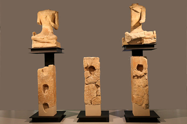A museum arrangement of two cross-legged statues on pedestals above three rectangular pillars. Everything is badly damaged. The pillars each have holes carved out of the front for something to be held. Both statues are headless. The left statue is missing its arms below the elbow. The right one is missing its entire left side. It has a torc around its neck.