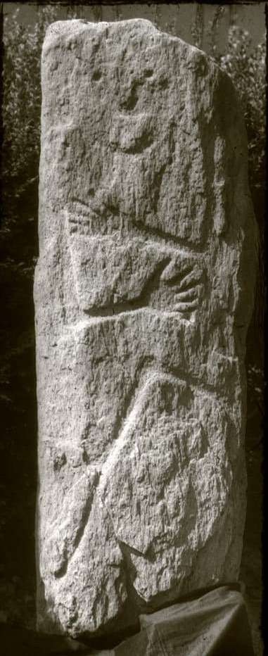 Old black and white photo of a natural looking pillar of stone with a roughly flat face on which has been inscribed the relief of a human figure. The eyes are small circles and the mouth looks like the curve of a smile. Its hands are in the same position as the Hirschlanden warrior, with splayed fingers. Beneath its hands is a sickle shaped tool. No other anatomy is drawn.