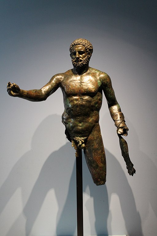 A dark gleaming bronze statue of Hercule, mounted on a pole, missing His right leg completely and the left leg below the knee. He is sculpted in life-like detail, with very curly short hair and a furrowed brow. His right hand reaches out while His left forearm is wrapped in a bandage that hangs down to his knee (although his left hand is missing).