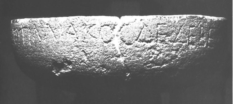 Black and white photo of the limestone basin inscribed with the votive, shown from the side. The inscription is rustic Greek capitals, the basin is very worn and pock marked and cracked. It is shaped like a bowl, but not quite as tall as a cereal bowl would be (proportionally speaking). It is 61 centimeters in diameter.