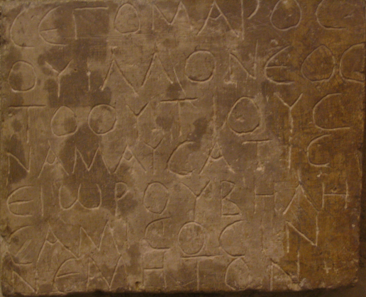 A color photo of the original inscription. The stone is a nearly-square rectangle, like a plaque, and its surface is brownish in color with an uneven patina. The text is written in rounded, rustic Greek capitals with thin lines that resemble handwriting with a stylus or pen.
