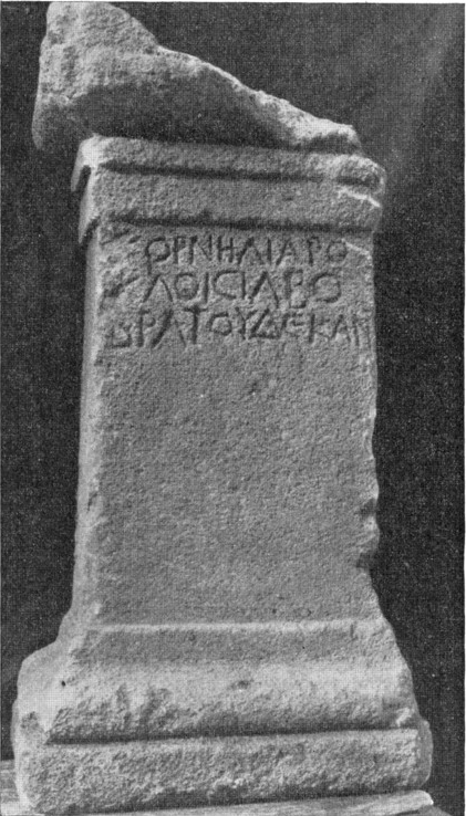 A black and white photo of a large, rectangular cippus, with a large base and crowning. The crowning's right corner has broken off. The inscription—in rustic Greek capitals—is borne on the cippus in a neat cluster towards the top, almost as if to save space, leaving the majority of the column looking blank. A ray of light shines down on the altar from above, giving it a holy aura.