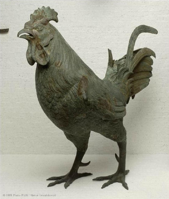 A color photo a bronze rooster with dark green and brown patina. It has an almost scientific anatomical detail, with fully realized plumage and even individual tail feathers sculpted separate from one another. It has an angry expression on its face with an open beak and is stepping forward like going after some unseen foe. Large talons and spurs on the back of its legs give it a vicious look.